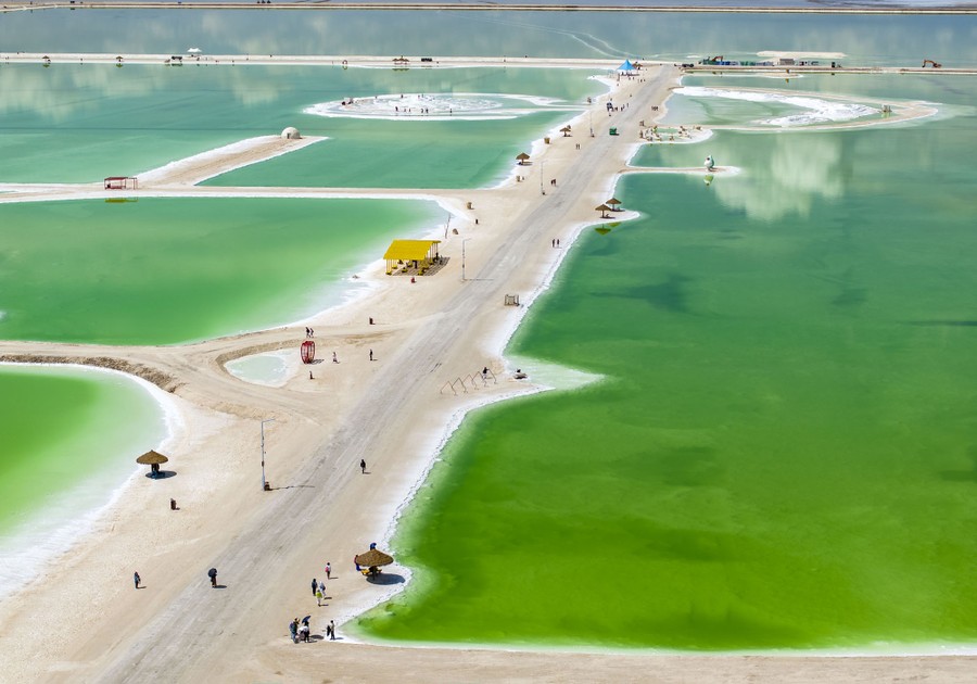 An aerial view of a long lane of sand through green-colored salt water in a shallow lake.