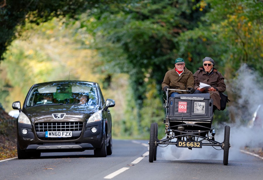 Two people drive a vintage steam-powered car next to a modern vehicle.