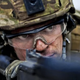 A person in camouflage clothing and helmet, wearing safety goggles, peers down the barrel of a gun.