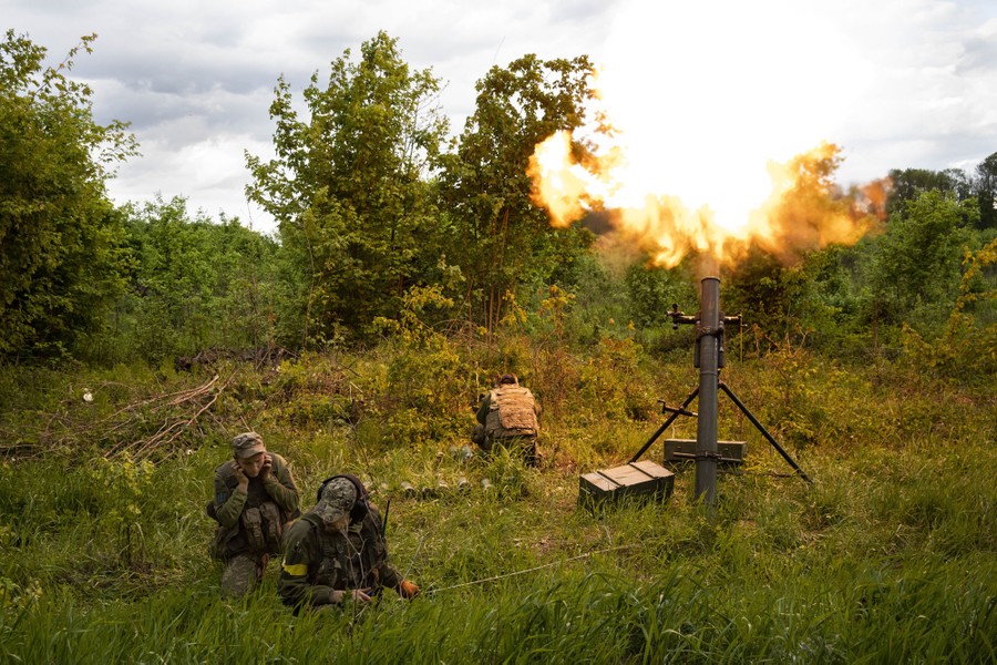 Three soldiers crouch beside a mortar launcher in a field as they fire a round.