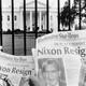 A black-and-white photo of two people reading newspapers that read “Nixon Resigning” outside of the White House gates