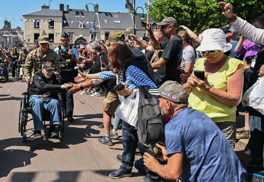 A crowd of onlookers cheer and reach out to shake the hand of an elderly American veteran being pushed in a wheelchair.