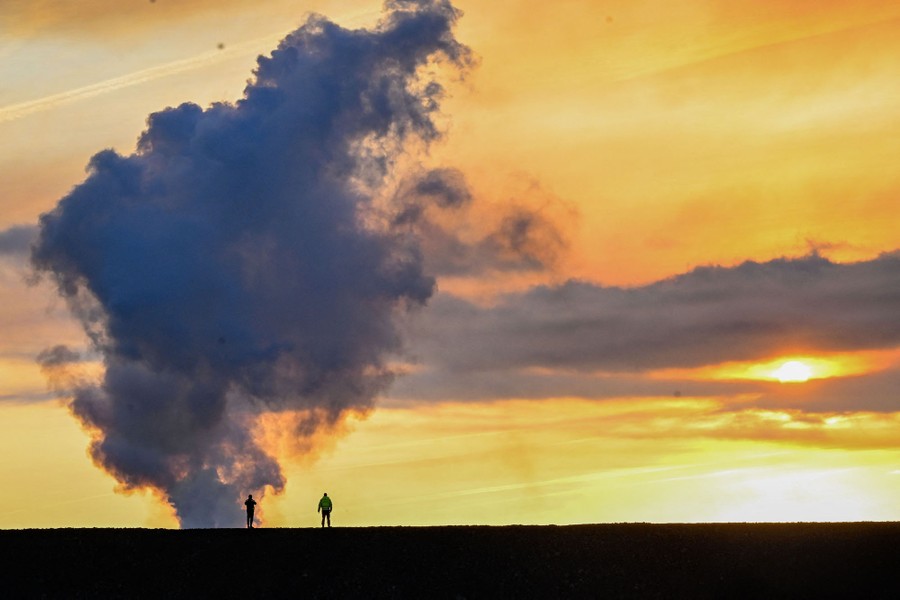 Two people stand on a flat landscape, silhouetted in front of a plume of volcanic gas, steam, and smoke.