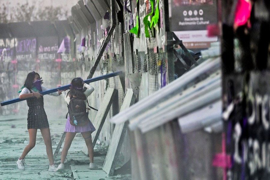 Two women use a pole to smash at a tall security fence during a protest.
