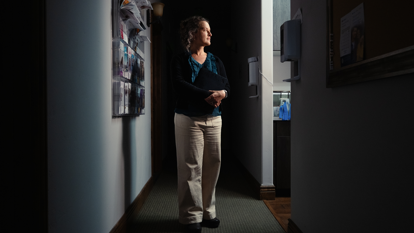 A photo of woman standing in shadowy hallway next to a patient exam room