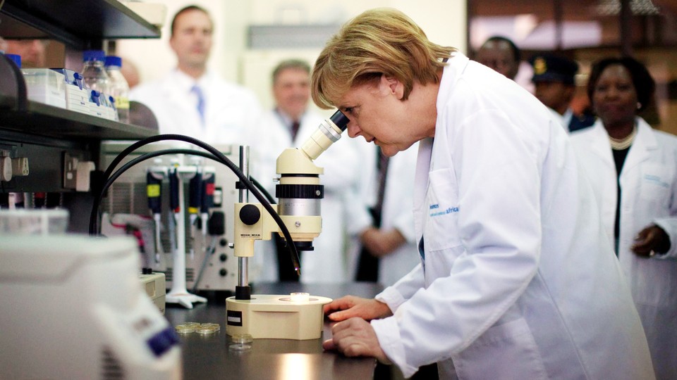 Angela Merkel looks through a microscope while wearing a white lab coat.