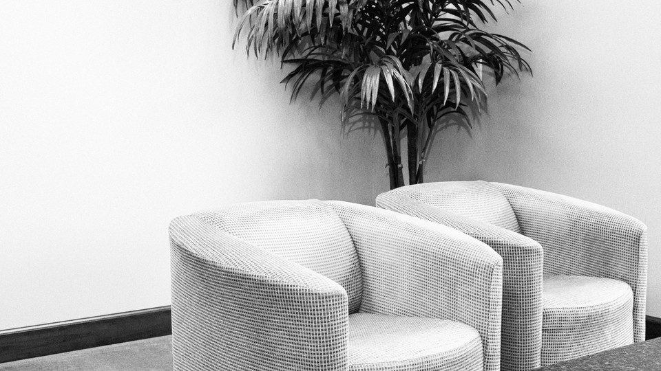 A black-and-white photo of a room with blank walls, two armchairs, and a potted plant