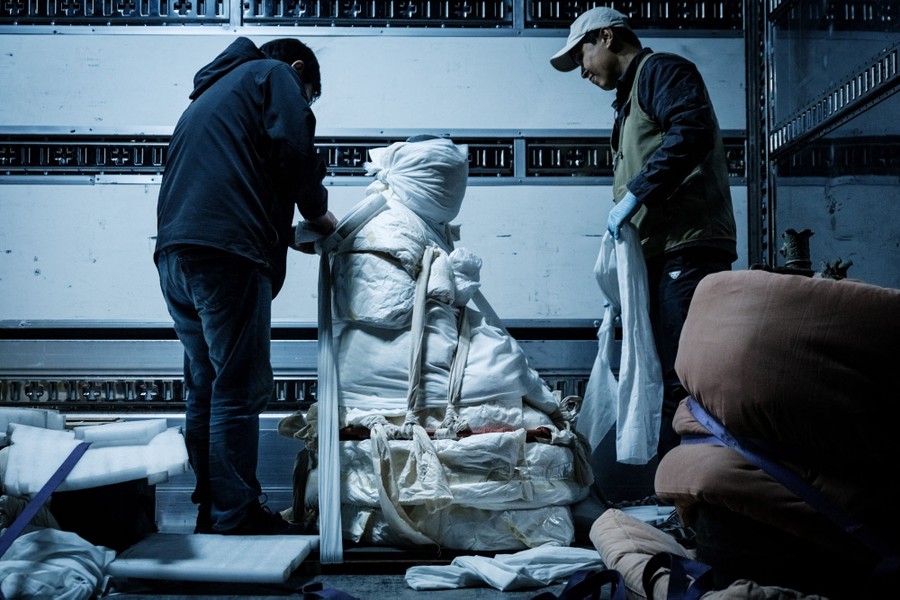 Two people work to wrap a Buddha statue that has been loaded into the back of a cargo truck.