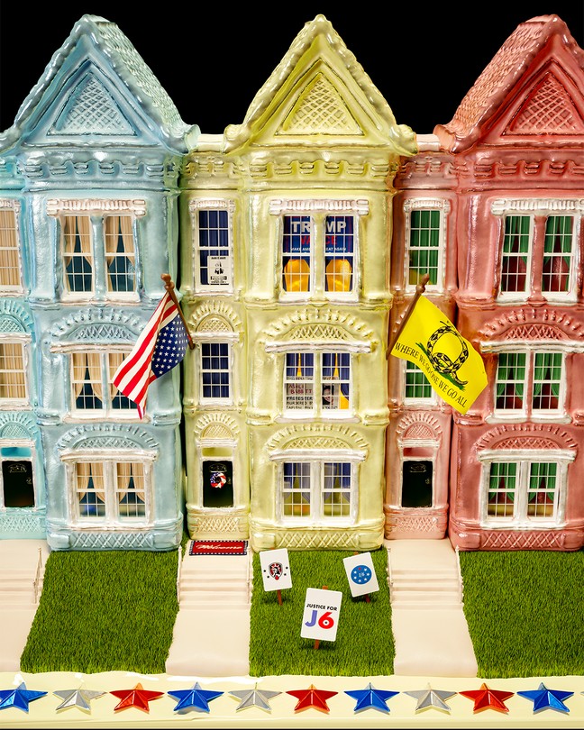 illustration with sculpture of 3 three-story rowhouses, blue, white, red, with green grass and front steps. The middle house has an upside-down U.S. flag and Q-Anon flag, yard signs about J6, Trump flag in window, and other signs.