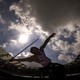 An athlete stretches and leans back while preparing to throw a javelin inside a stadium, silhouetted against a cloudy sky.