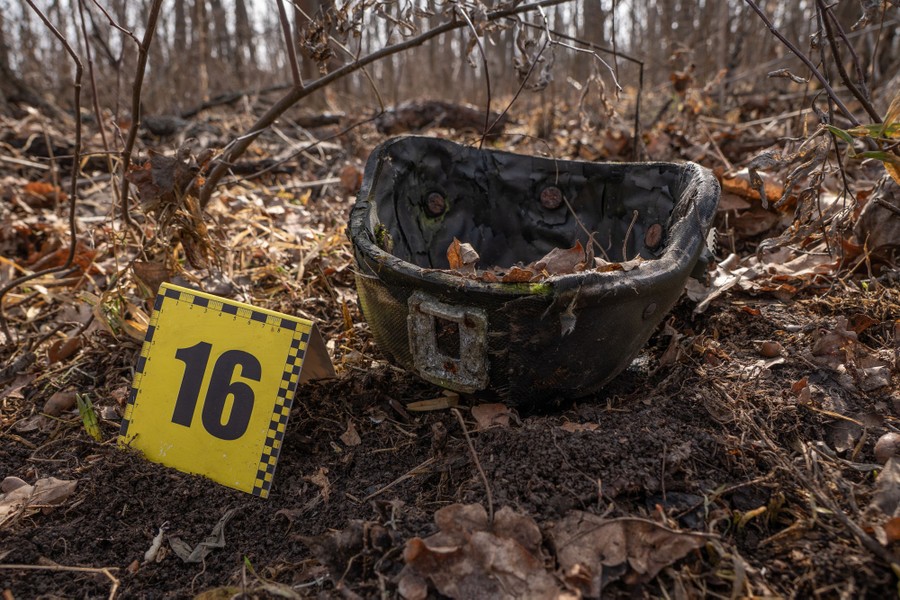 A numbered evidence card sits on the ground beside an empty dirt-and-leaf-covered military helmet in a forest.