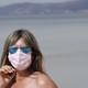 A woman on a beach in blue reflective sunglasses and a pink face mask