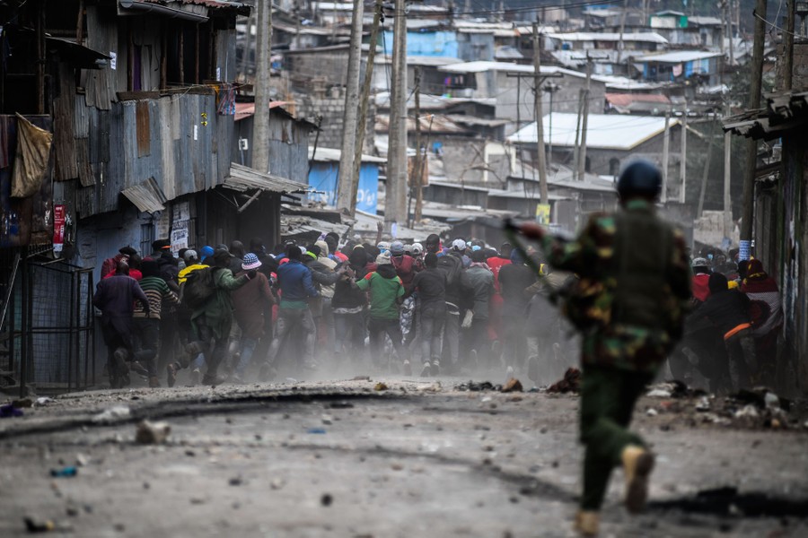 A riot-police officer runs toward a large crowd of fleeing protesters in a street.