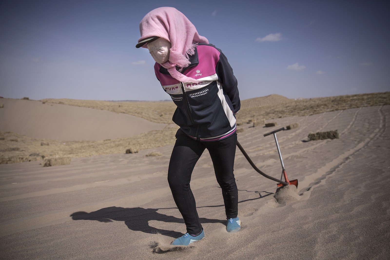 A woman pulls a small shovel behind her, plowing a shallow trench in sand.