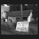 Black and white photo of ruined house with hand-painted sign "The System Is the Problem"