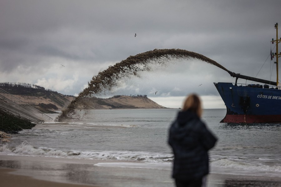A person on a beach watches as a nearby ship sprays silt toward the shoreline.