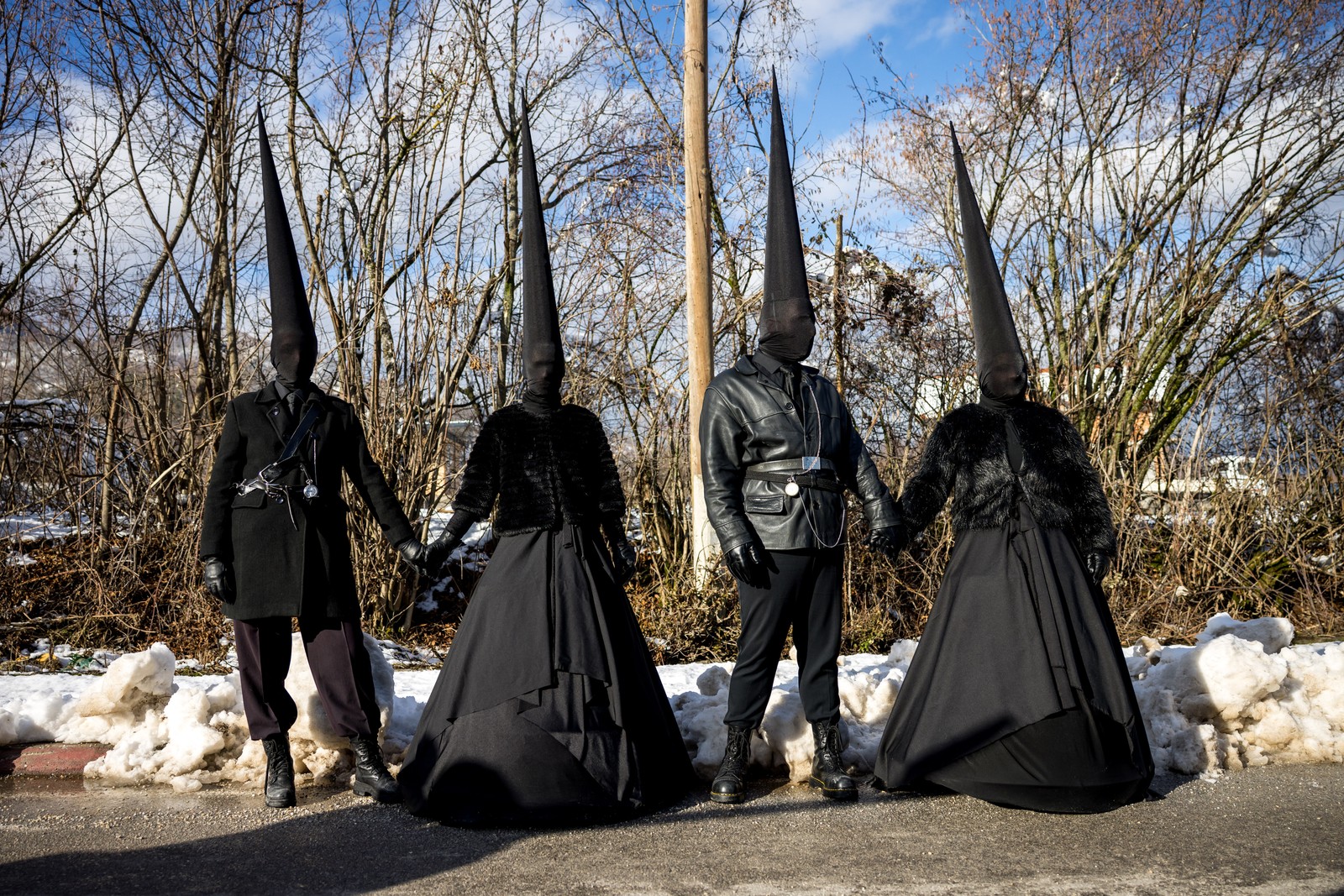 Two couples pose, all wearing costumes of black clothing, with tall conical black caps and black cloth over their heads.