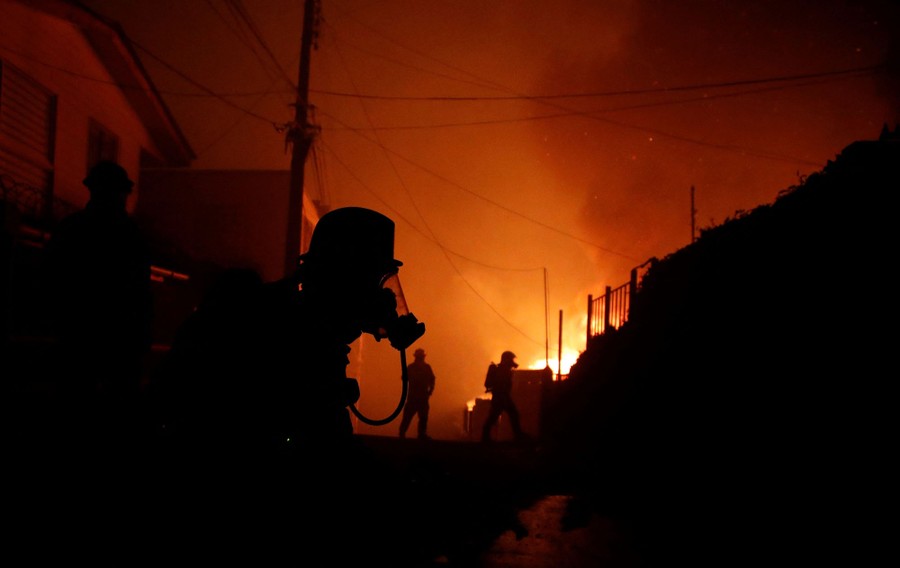 Several firefighter work in a street, backlit by a forest fire.