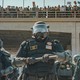 Police officers stand in a line in front of a bridge with people standing on it