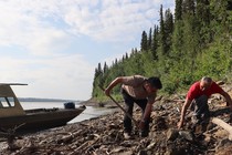 A photograph of two men digging for fossils along the Alaskan Yukon River, with a boat docked behind them