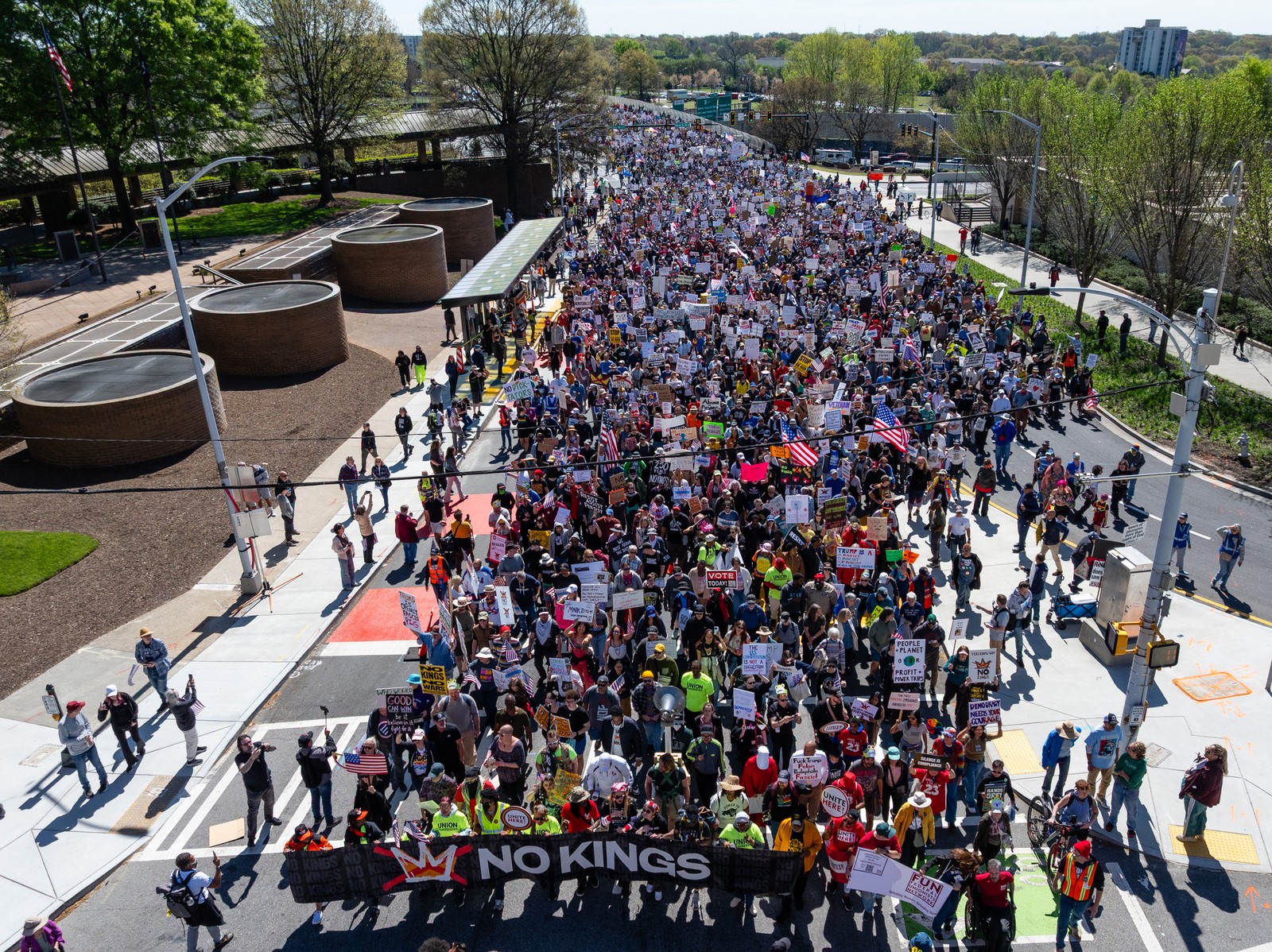 A group of thousands of protesters march down a city street, behind a banner carried at front, which reads 