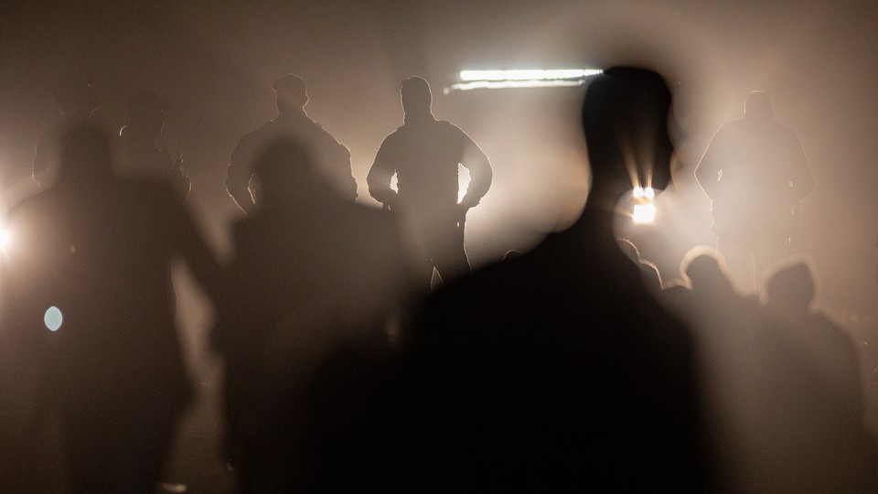 Photograph of black silhouettes of border agents standing with hands on their hips illuminated by a vehicle’s headlights behind them