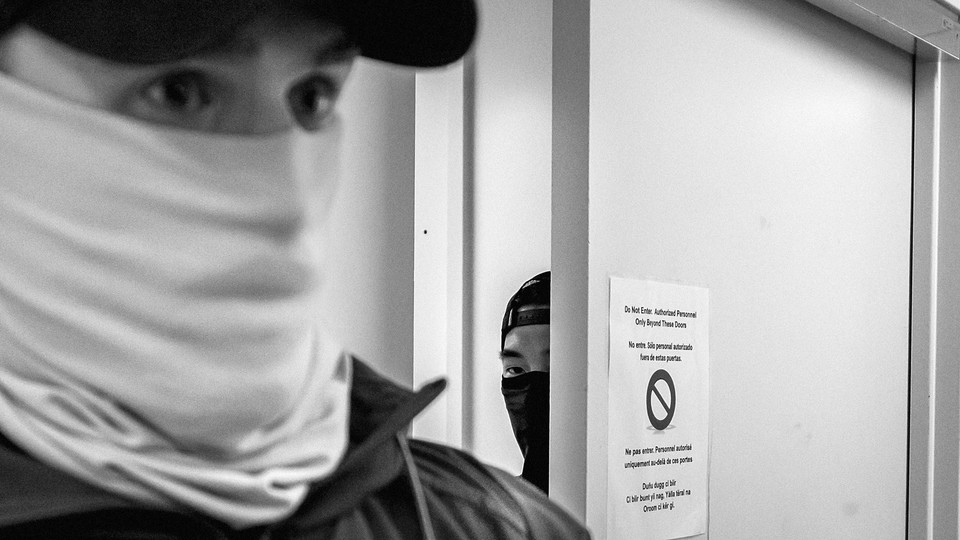 A black and white photographs of two ICE Officers, one in the foreground, and one peeking out from behind a door.