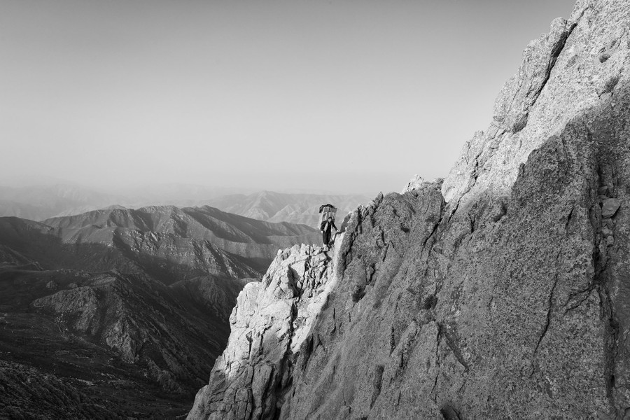 A person carries a heavy pack of materials on their back as they walk across bare rock on the side of a steep mountain.