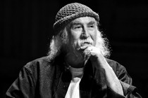 a black and white image of David Crosby in a knit cap with his hand to his chin