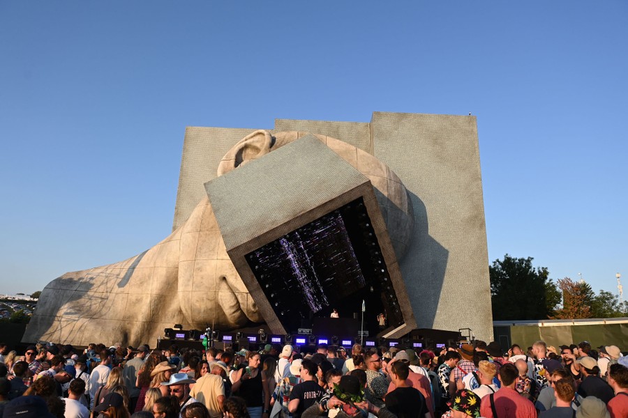 A crowd stands before a stage that is designed to look like a gigantic human head with a large rectangular visor.