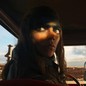 A film still showing Anya Taylor-Joy as the character Furiosa looking backward while sitting in a car; other cars, and an explosion, can be seen through the windshield in the desert landscape behind her.