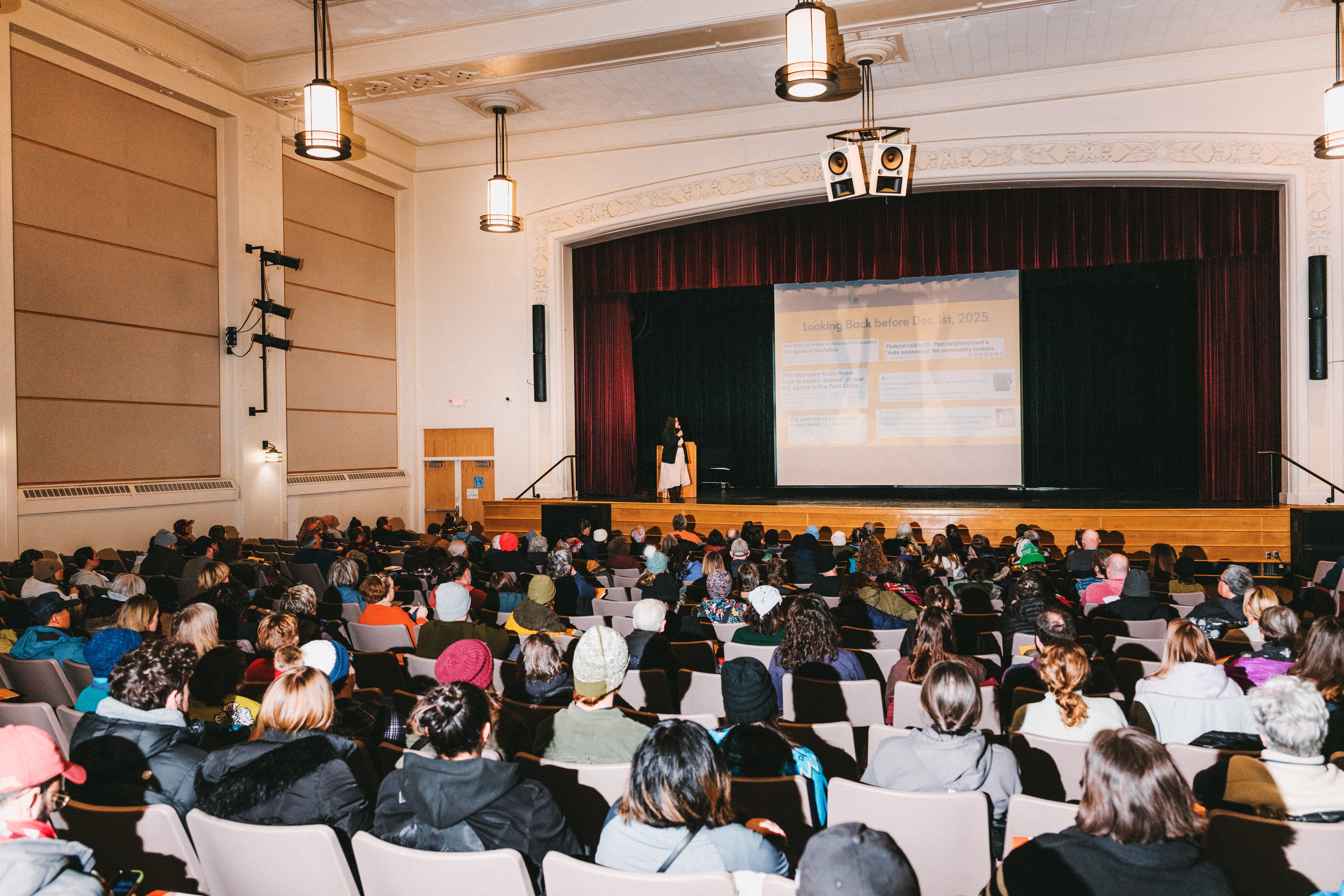 A person on a stage in an auditorium is seen in front of a large seated crowd