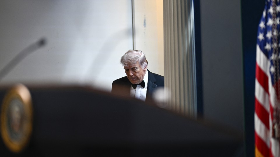 Trump in tuxedo entering White House briefing room; American flag visible on the side and podium in the foreground