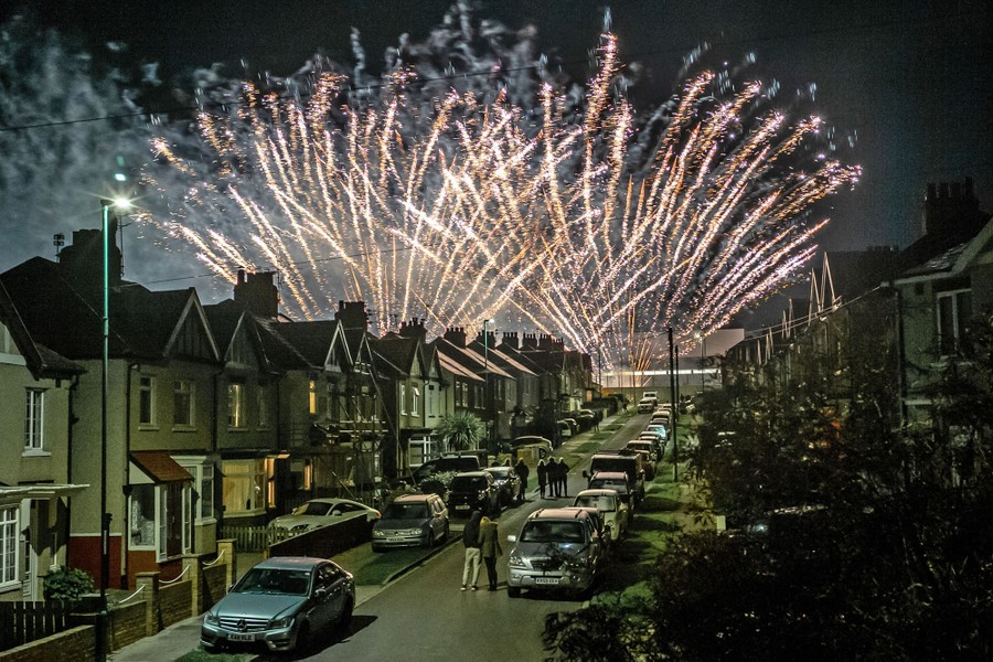 People stand in a residential street, watching fireworks erupt above houses.