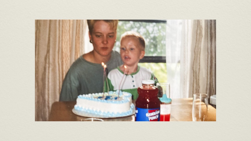 A photograph of the author as a young child, with close-cropped blond hair and wearing a green-and-white henley, on the lap of his mother, sitting in front of a blue and white heart-shaped cake with three lit candles in it