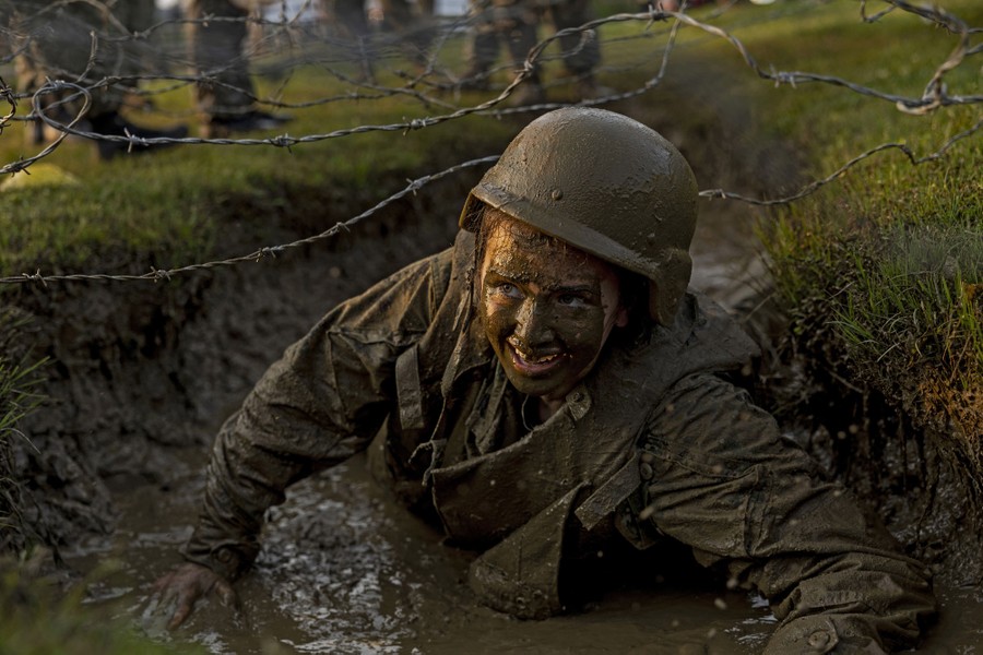 A person in military gear crawls through a muddy trench under barbed wire.