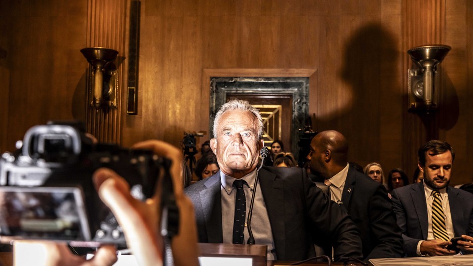 A color photo of RFK Jr. in a suit, in a wood-paneled room, surrounded by other people in suits and lit dramatically by a camera flash