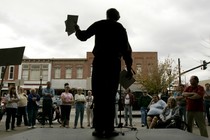 Al Norman, seen in silhouette, holds flyers and speaks to a crowd from a stage.