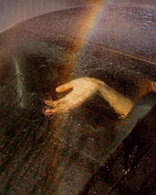A hand, palm facing upward, reaches for the glass of the back window of a car. Rain falls onto the surface of the car, and a rainbow cuts through the center, over the hand.