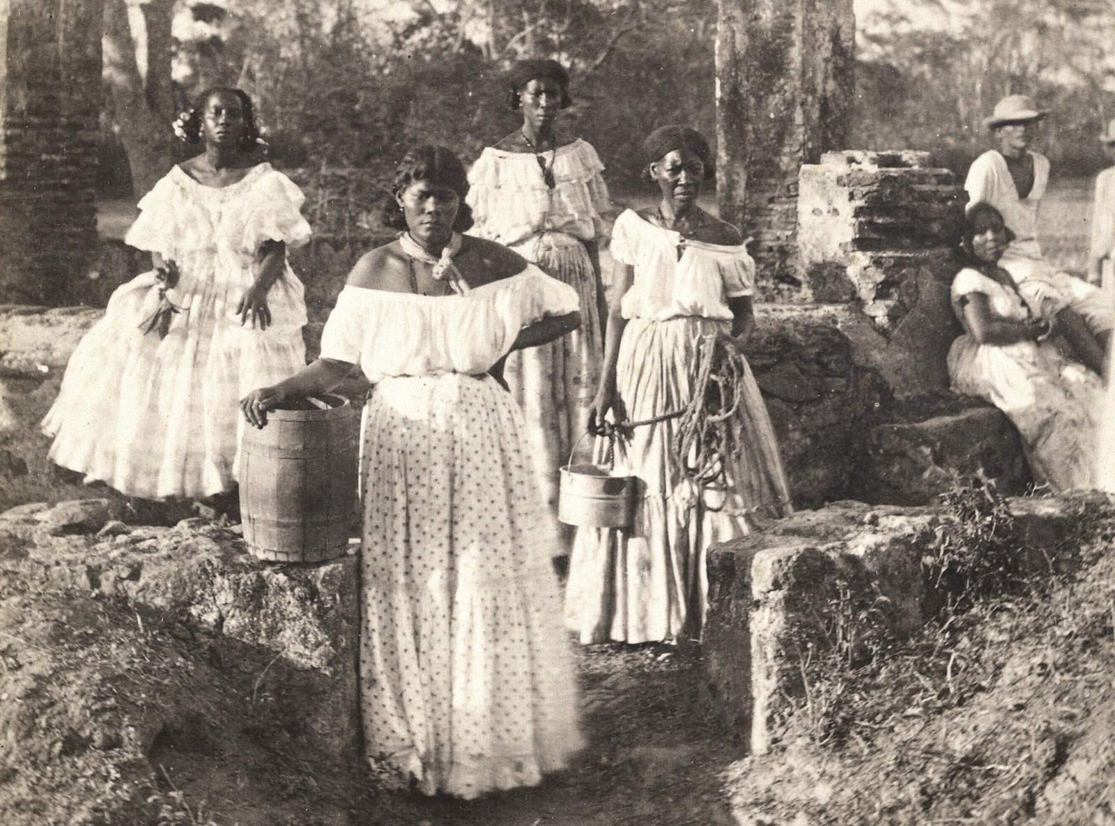Five women and a couple of men sit and stand outside, posing in white clothing. One holds a bucket and long rope.