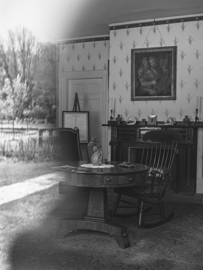 black-and-white photo of antique-furnished interior of room with table and fireplace, and partial reflection of outdoor garden and path