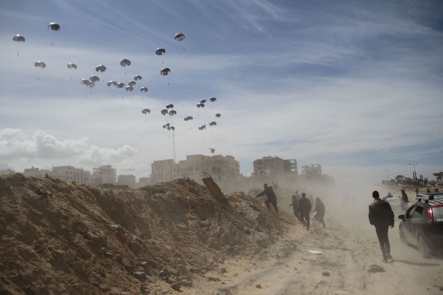 People on a dirt road look up toward parachutes falling toward distant buildings.