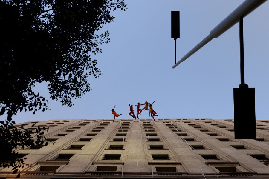 Six dancers perform on the side of a building, hanging from ropes.