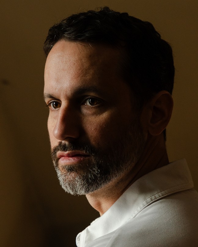 A white man wearing a white shirt stands in front of a yellow wall