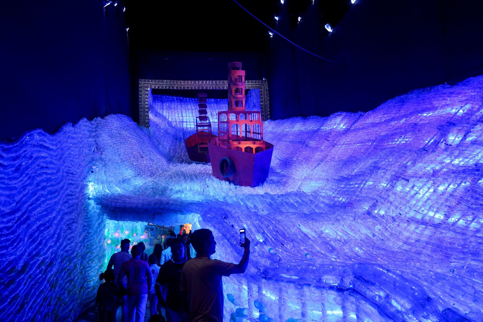 A person takes a photograph of a large sculpture made of plastic, shaped like ocean waves with boats on it.
