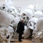 A person stands in an exhibit space amid a large pile of giant human-skull sculptures.