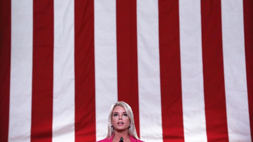 A color photo of Pam Bondi speaking in front of a large, vertical American flag.