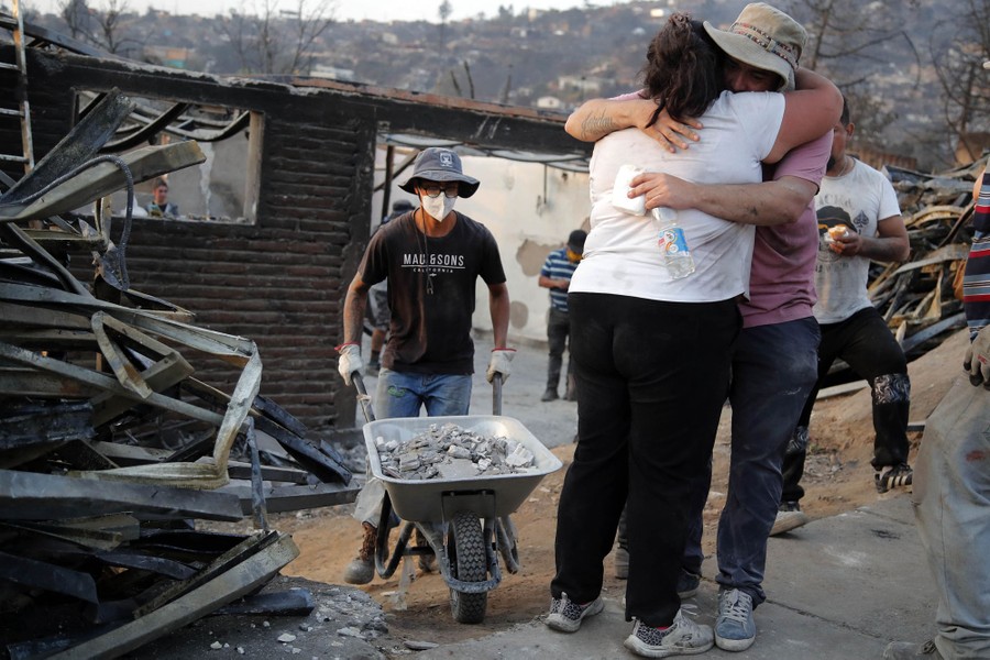 Two people embrace as others work to clear debris from a burned house.