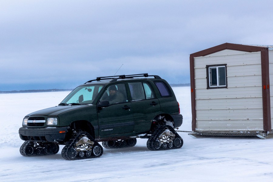A car, with triangular caterpillar tracks where its wheels would normally be, drags a small shack behind it on an ice road.