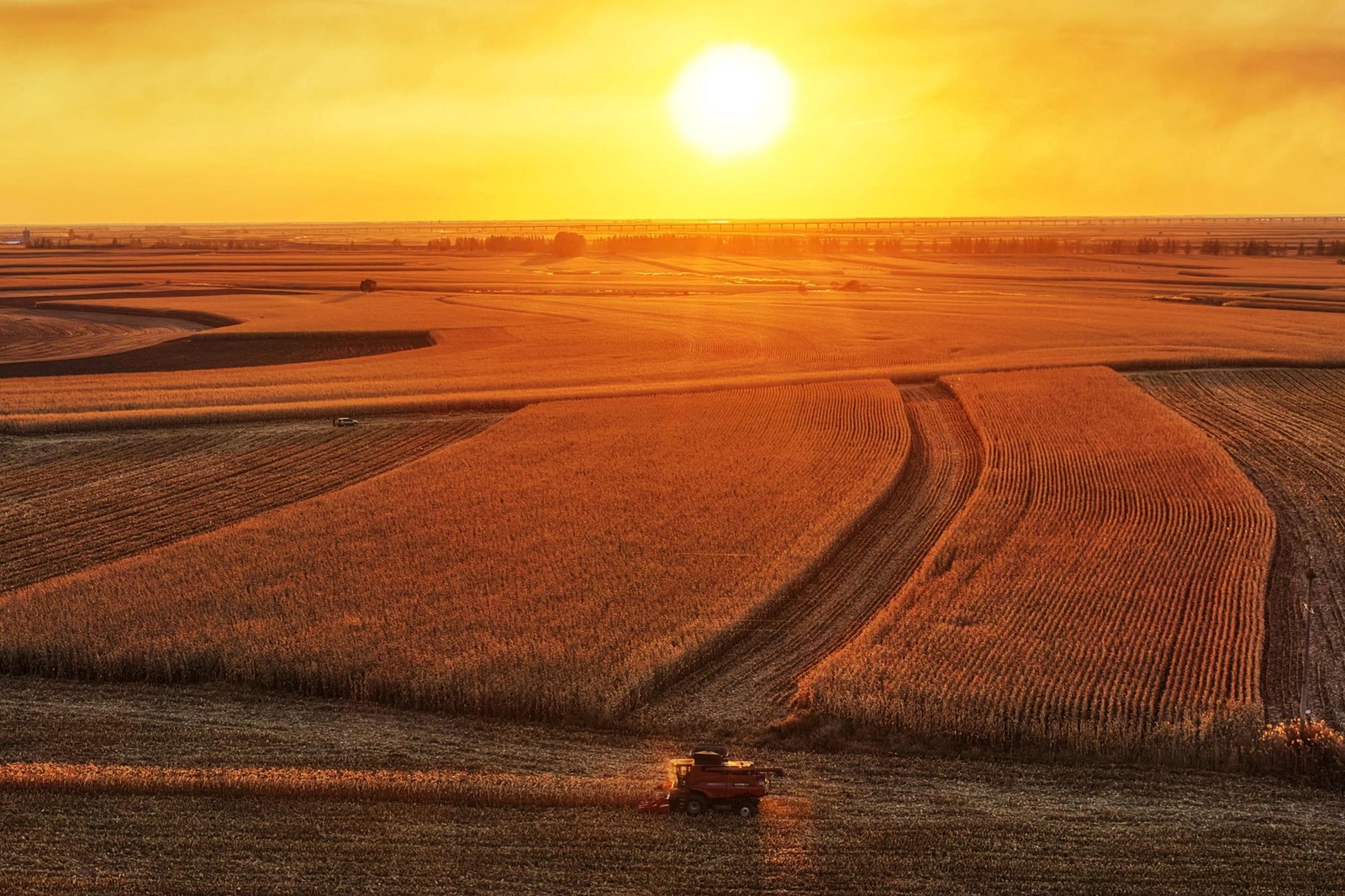 An aerial view of a piece of farm equipment being used to harvest grain from a field, seen in a broad landscape of farm fields, at sunset